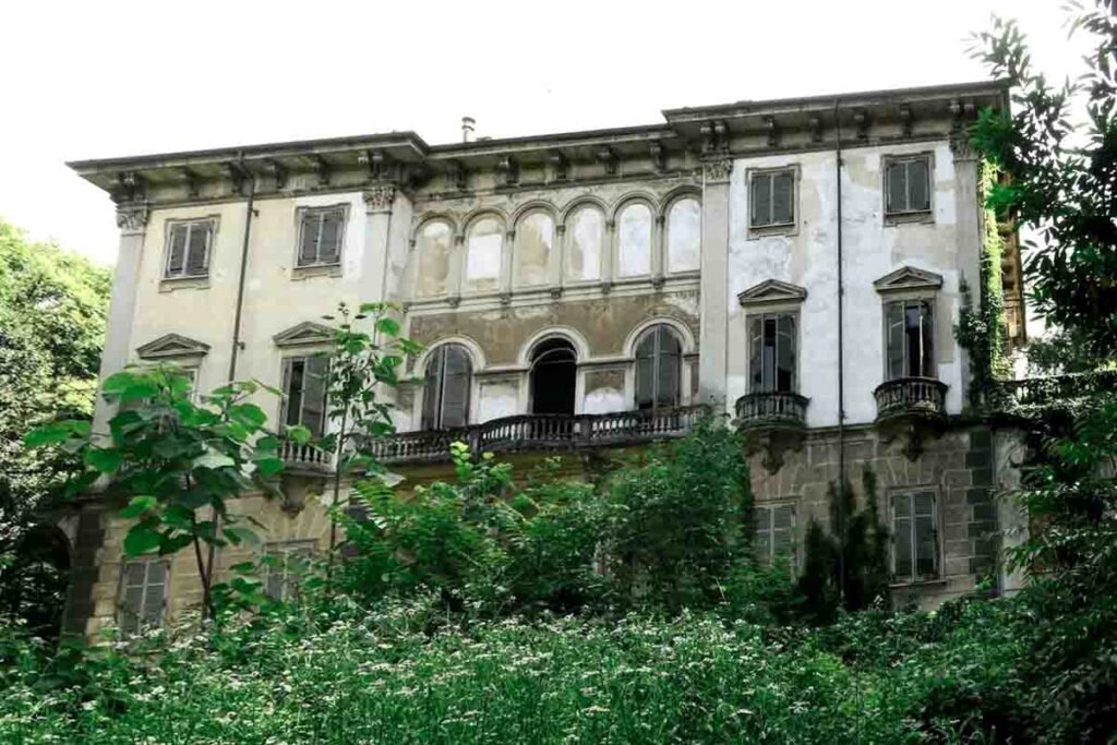 Exterior facade of a large, historic abandoned villa surrounded by overgrown plants and shrubs.